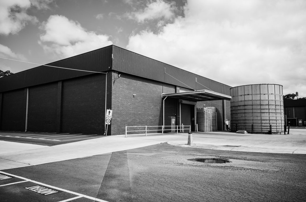 Black and white photo of a large industrial warehouse with a storage tank and parking area.